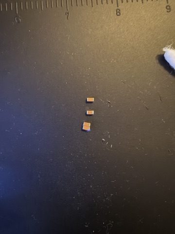 Three tiny rectangular ceramic capacitors sitting on a dark mat next to a ruler.