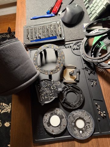 A first-generation HomePod taken apart on a black repair mat, with the speaker driver rings, the boards, the screws and a precision bit kit laid out around it.