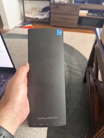 A hand holding a slim black Dell OptiPlex 3090 Ultra upright in a living room.